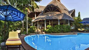 A swimming pool with lounge chairs and umbrellas, surrounded by tropical vegetation and a thatched-roof building. - Large luxury beachside villa with a pool, guesthouse, cook, gardener, and maid (Diani Beach)
