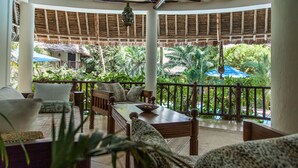 A covered outdoor seating area with a wooden table and chairs, surrounded by lush greenery and a thatched-roof structure in the background. - Large luxury beachside villa with a pool, guesthouse, cook, gardener, and maid (Diani Beach)