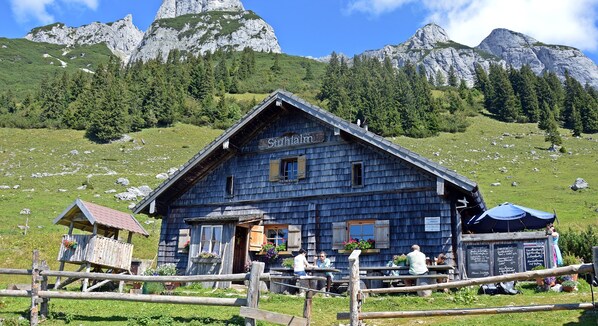 Un edificio de madera rústico con el letrero "Stuhlalm", una cerca de madera en primer plano y un paisaje montañoso que lo rodea.