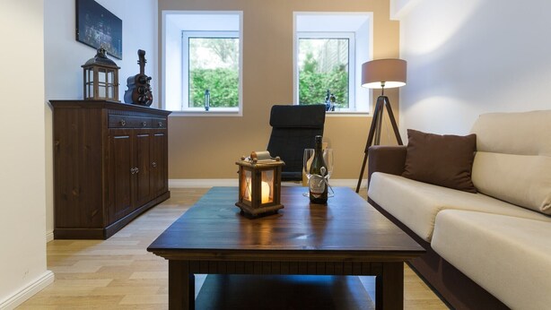 A living room with a dark wooden coffee table, a beige sofa, a dark wooden cabinet, and a lamp. There are two windows with a view of greenery outside. - with underfloor heating and WiFi (Berlin)