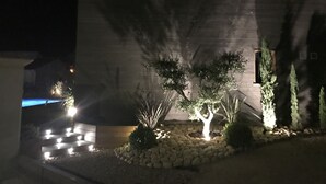 A nighttime view of a building with a well-lit garden area featuring rocks, shrubs, and a small tree. - Luxury villa with private pool 500m from the beach St Cyprien Porto Vecchio (Lecci)