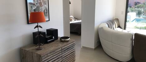 Una sala de estar moderna con un sofá, una consola de madera con dispositivos electrónicos, una obra de arte enmarcada y vistas al exterior a través de un ventanal.