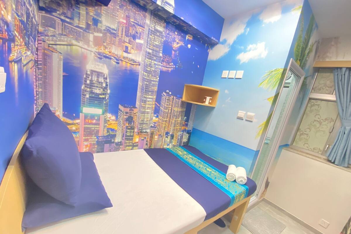 A room with a bed, a small shelf, and a mural of a cityscape.