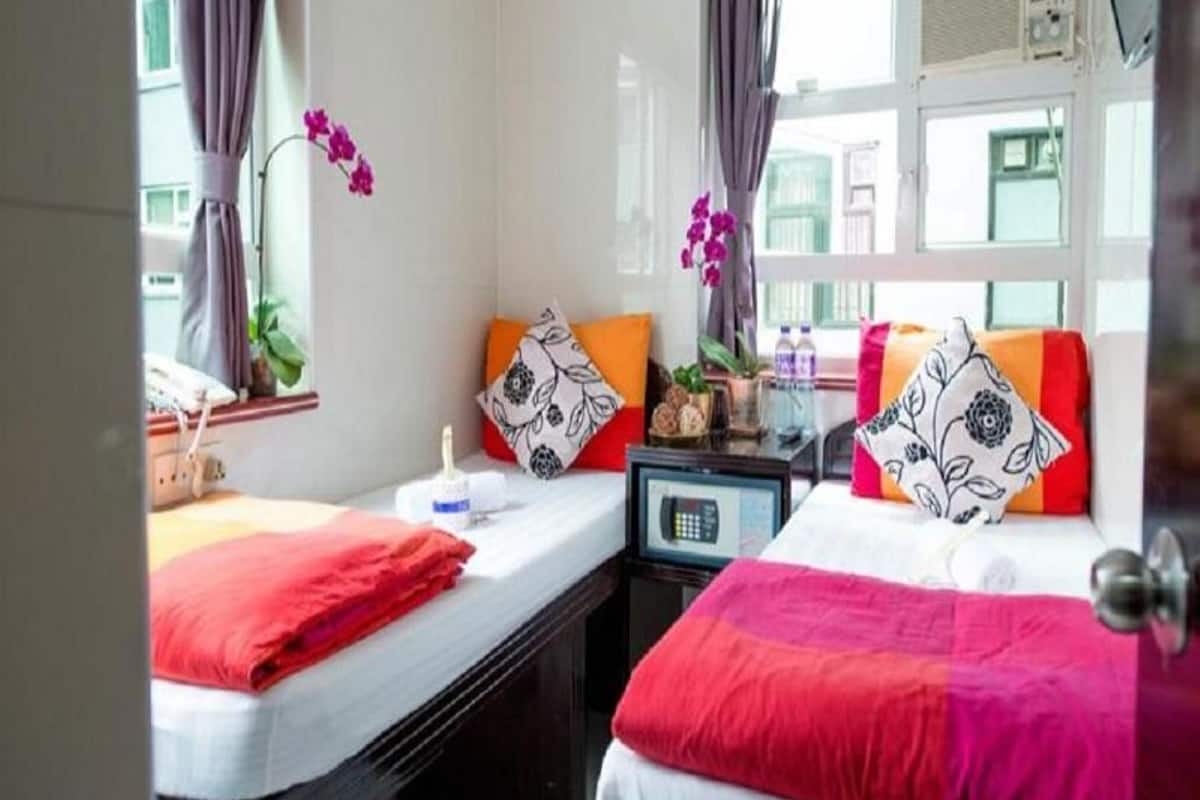A hotel room with two beds, each with a red blanket, a small table with a lamp, and a window with curtains.