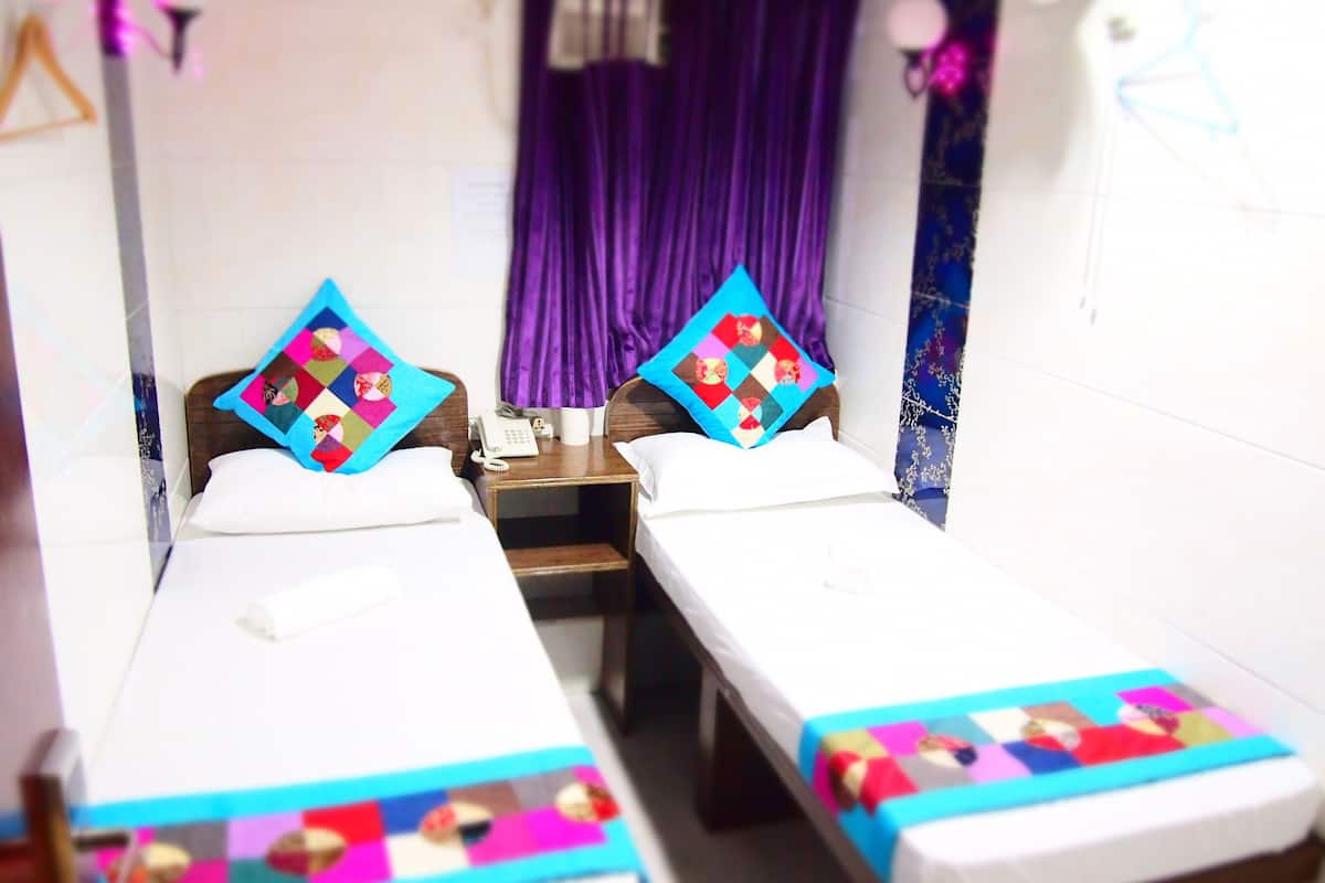 A hotel room with two single beds, each with a colorful quilt and a pillow. There is a wooden nightstand between the beds with a phone and a lamp. The room has purple curtains and a white wall with a decorative item hanging on it.