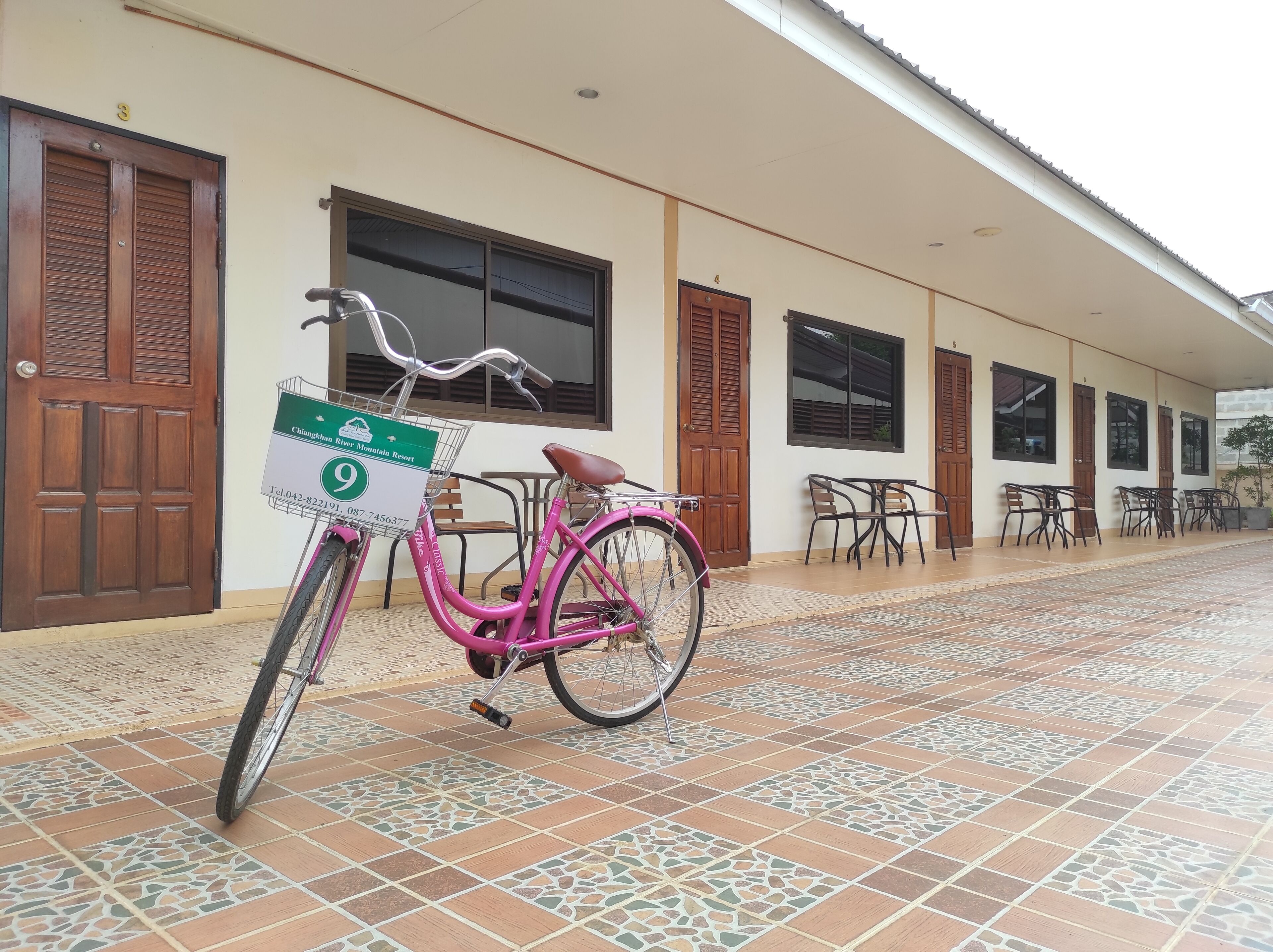 A pink bicycle parked in front of a building with numbered doors and a tiled patio area.