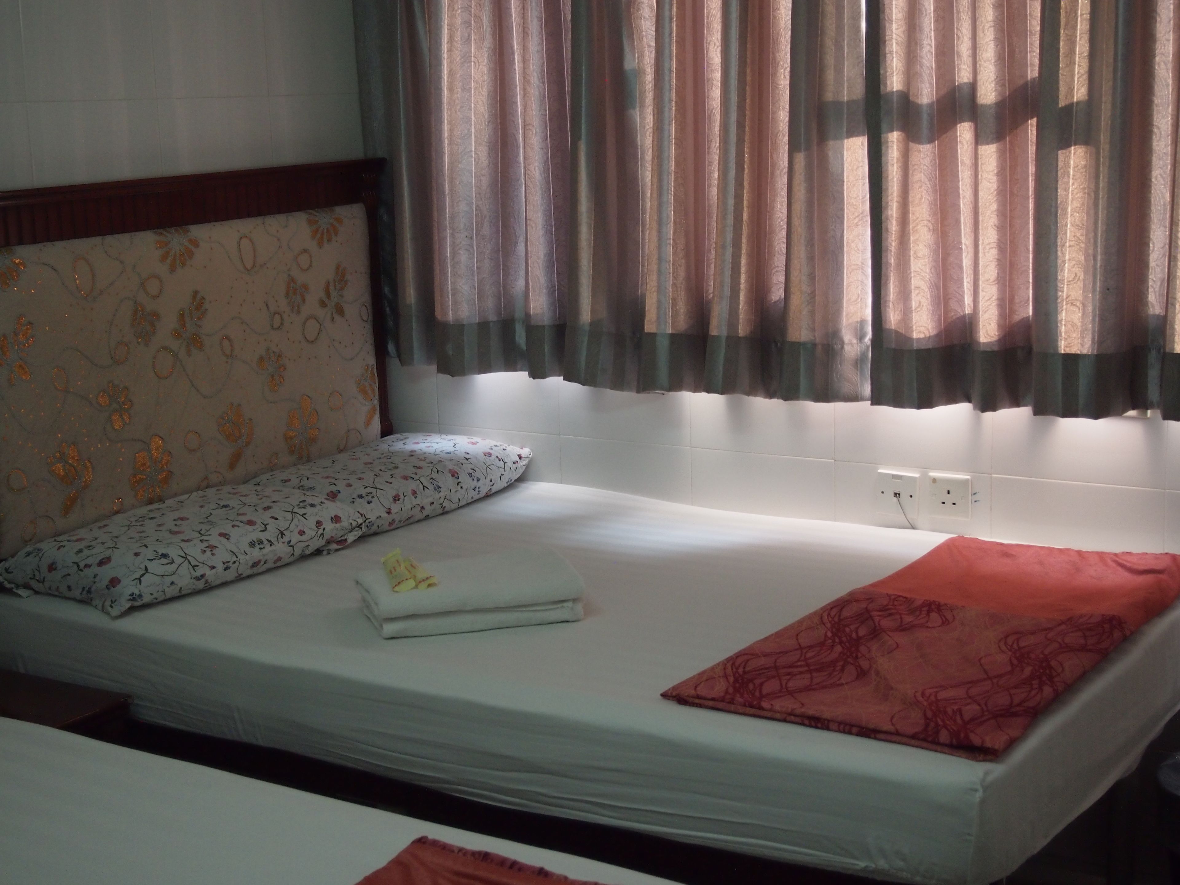 A single bed with a floral headboard, a bedside table with a folded towel, and a window with patterned curtains.