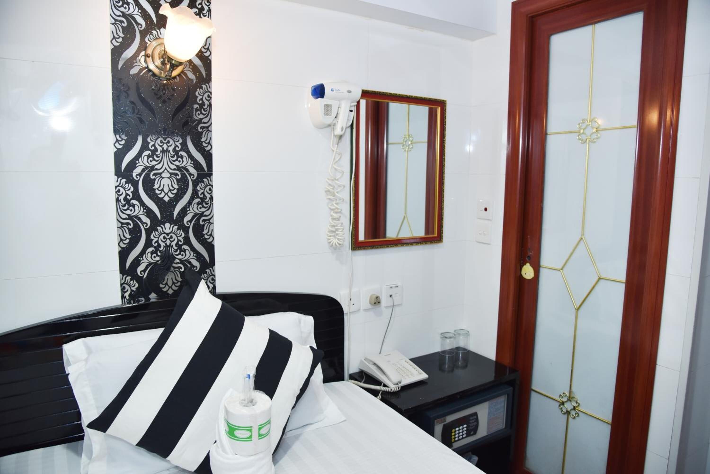 Una habitación de hotel con cama, mesita de noche con teléfono y gafas, espejo, secador de pelo en la pared y puerta con diseño geométrico.