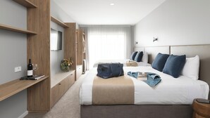 A modern hotel room with a large bed, a TV mounted on the wall, a wardrobe, and a bedside table with a wine bottle and glasses.