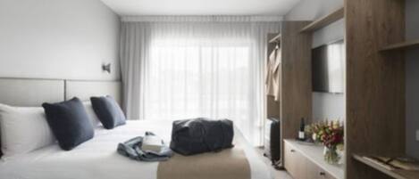 Una habitación de hotel moderna con una cama grande, un televisor de pantalla plana, un armario de madera y un ventanal con cortinas traslúcidas.
