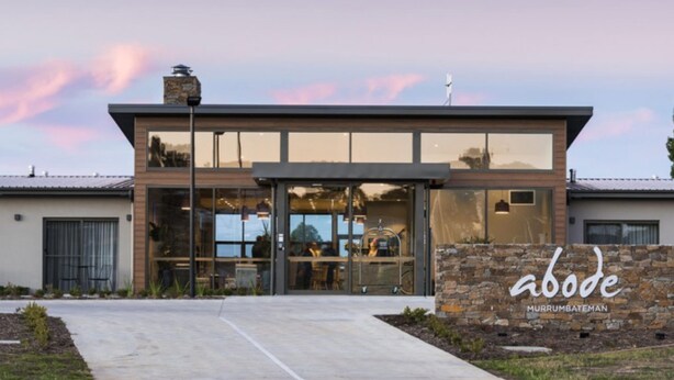 A modern building with a large glass facade, a stone wall with the name 'abode', and a clear sky.
