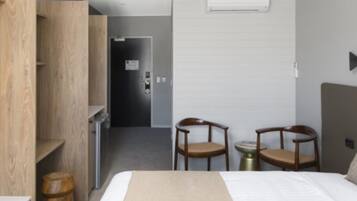 A hotel room with a bed, wooden wardrobe, two chairs, a small table, and a door leading to a hallway.