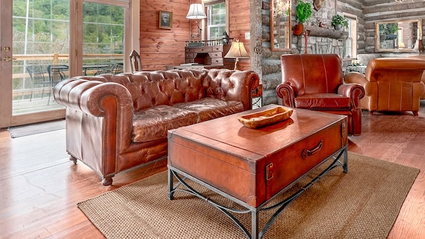 A log cabin interior with a leather sofa, a wooden coffee table, and a stone fireplace. - TWO Amazing Log Cabins, Storybook Setting, Hot Tub, WiFi, Mountain Views Pets Ok (Gerton)