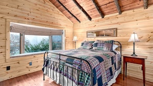 A cozy bedroom with a wooden ceiling, a large window, a bed with a plaid bedspread, a bedside table with a lamp, and a framed picture on the wall. - TWO Amazing Log Cabins, Storybook Setting, Hot Tub, WiFi, Mountain Views Pets Ok (Gerton)