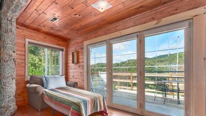 A wooden cabin room with a bed, a sofa, and large sliding glass doors leading to an outdoor deck. - TWO Amazing Log Cabins, Storybook Setting, Hot Tub, WiFi, Mountain Views Pets Ok (Gerton)
