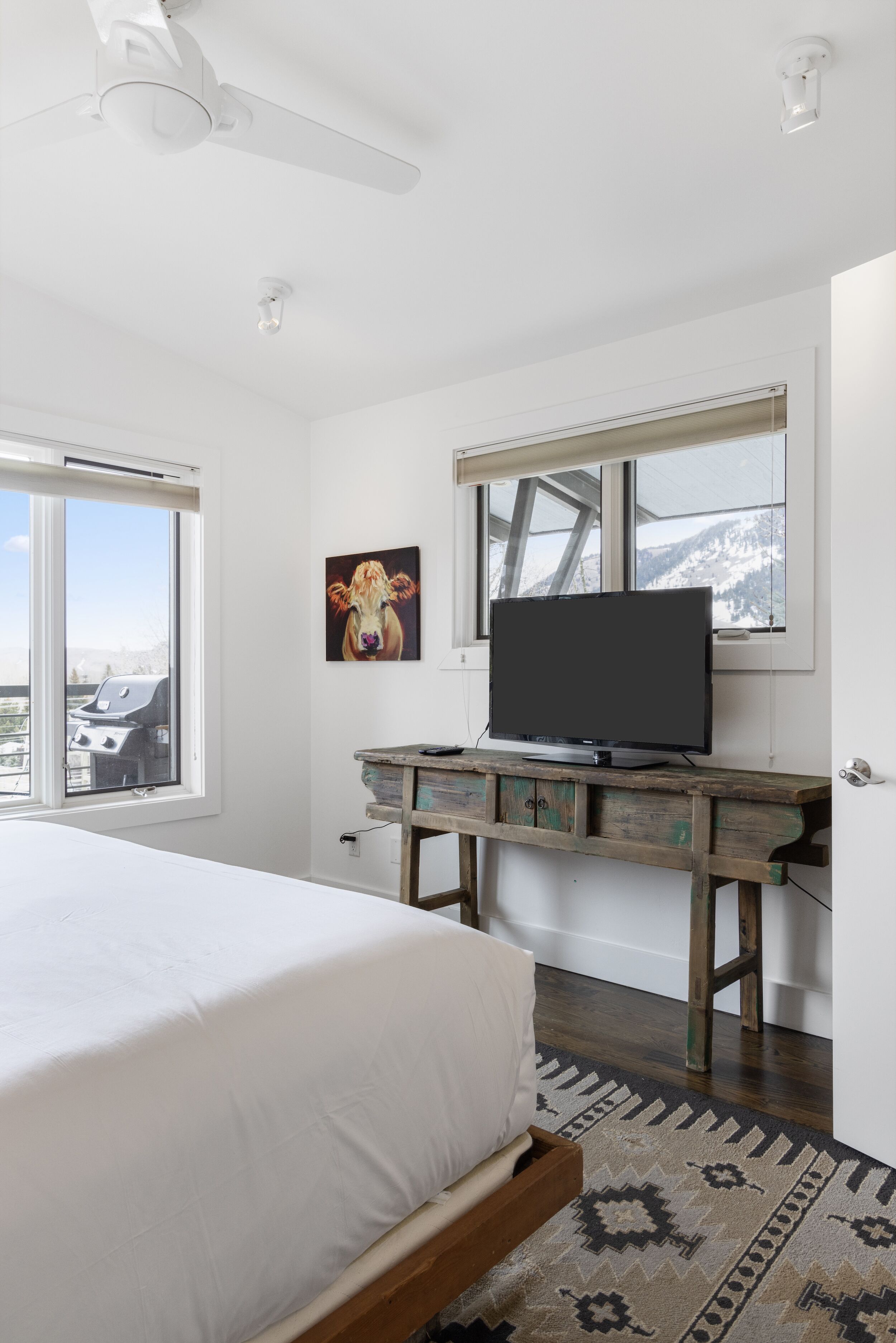 A bedroom with a bed, a television on a wooden dresser, a painting of a cow, and a window with a view of mountains.