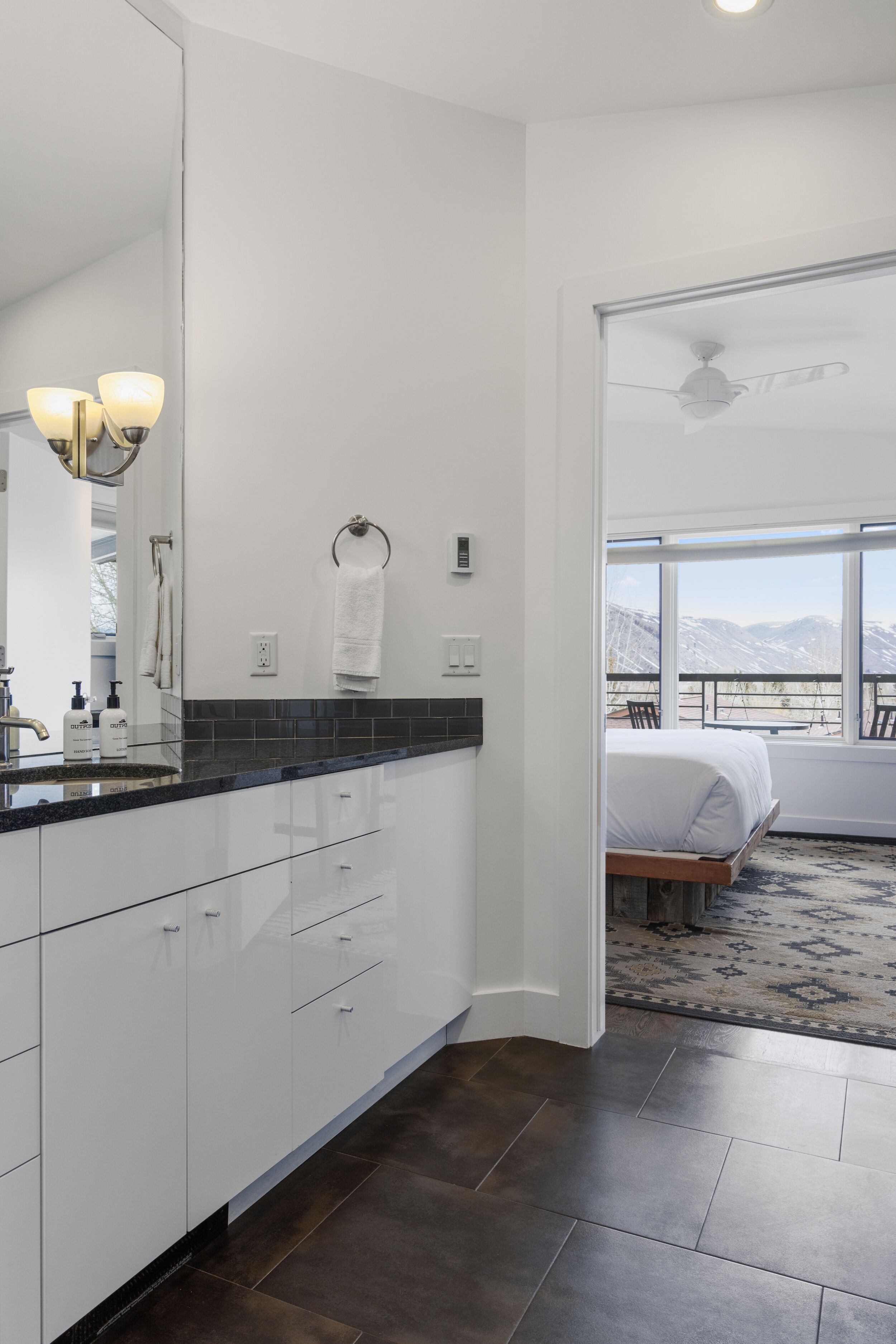 Une salle de bain moderne avec un grand miroir, des meubles blancs et un plan de travail noir. Une fenêtre offre une vue sur les montagnes.