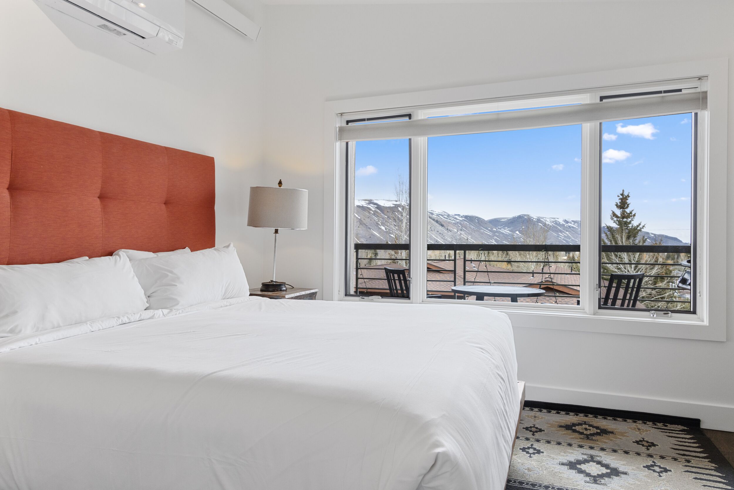 Habitación de hotel con una cama grande, un cabecero, una mesita de noche con lámpara y vistas a montañas a través de amplios ventanales.
