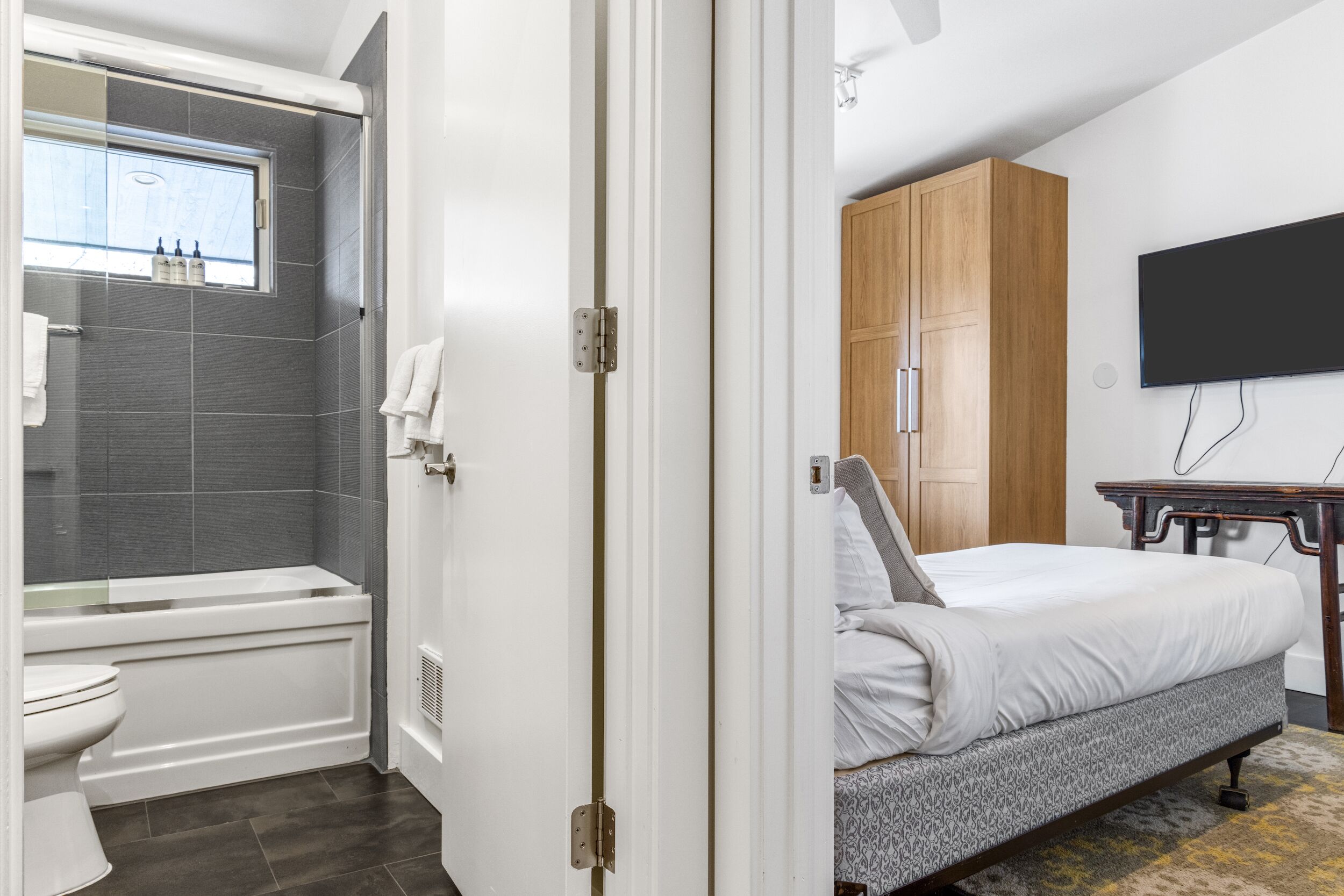 A modern bathroom with a bathtub, toilet, and shower. There is a wooden wardrobe and a bed with a patterned bedspread.