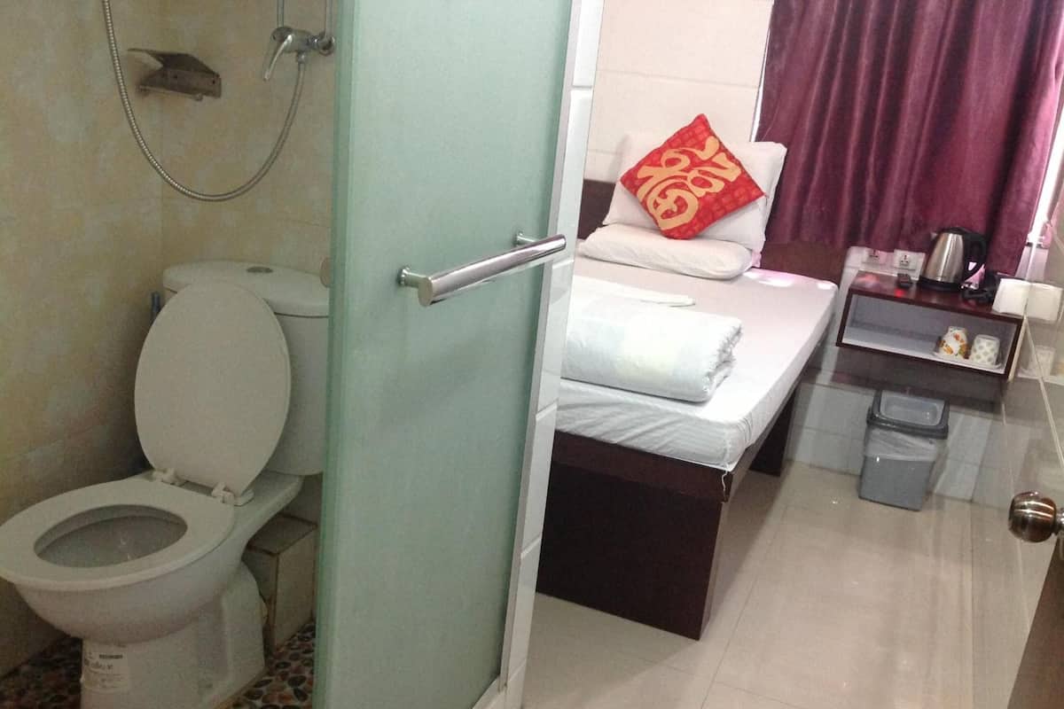 A bathroom with a toilet, a shower, a bed with a red pillow, and a bedside table with a kettle and a cup.