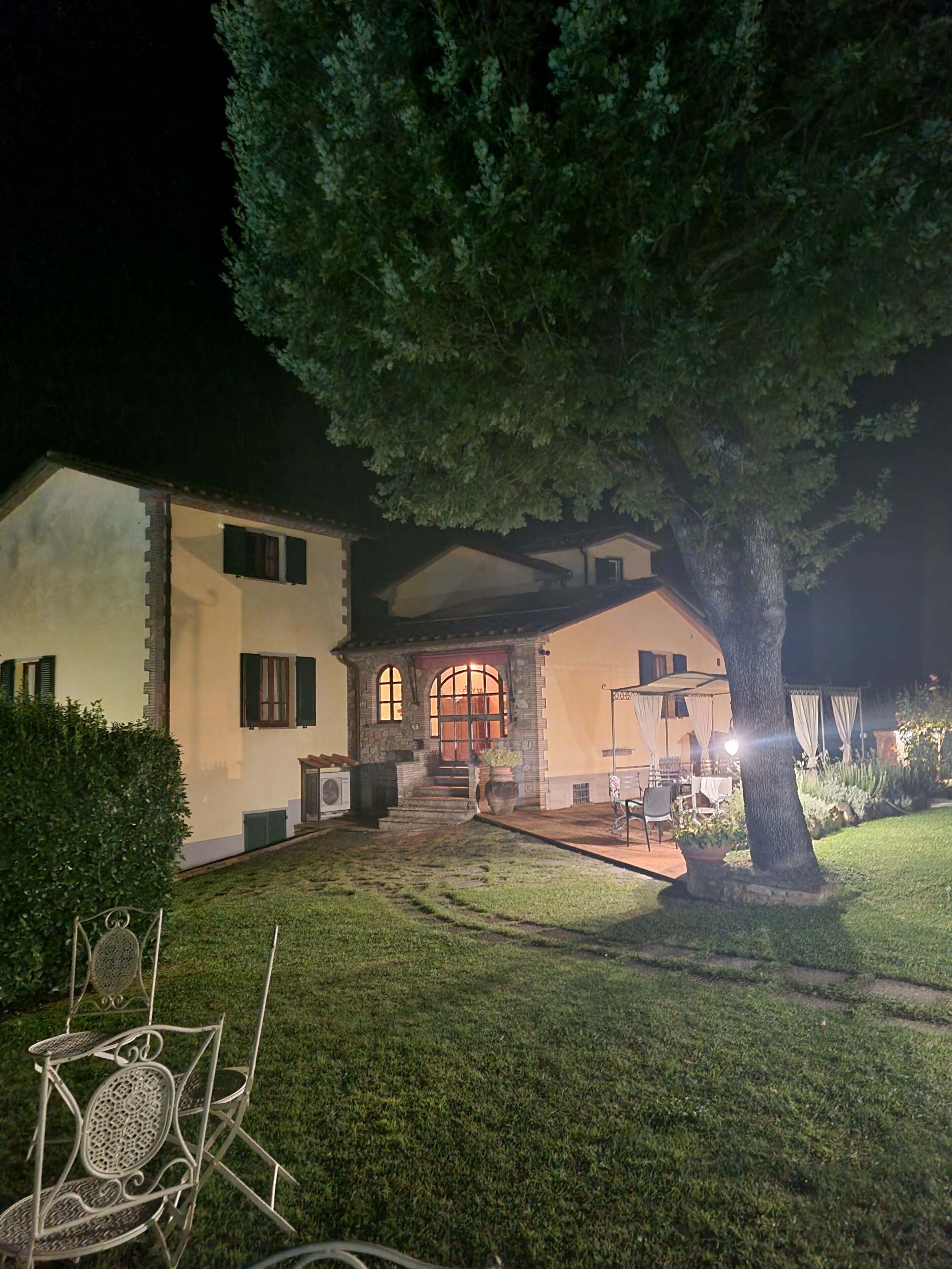 Une maison à deux étages avec une façade en pierre, un grand arbre à l’avant et un patio aménagé avec du mobilier d’extérieur.