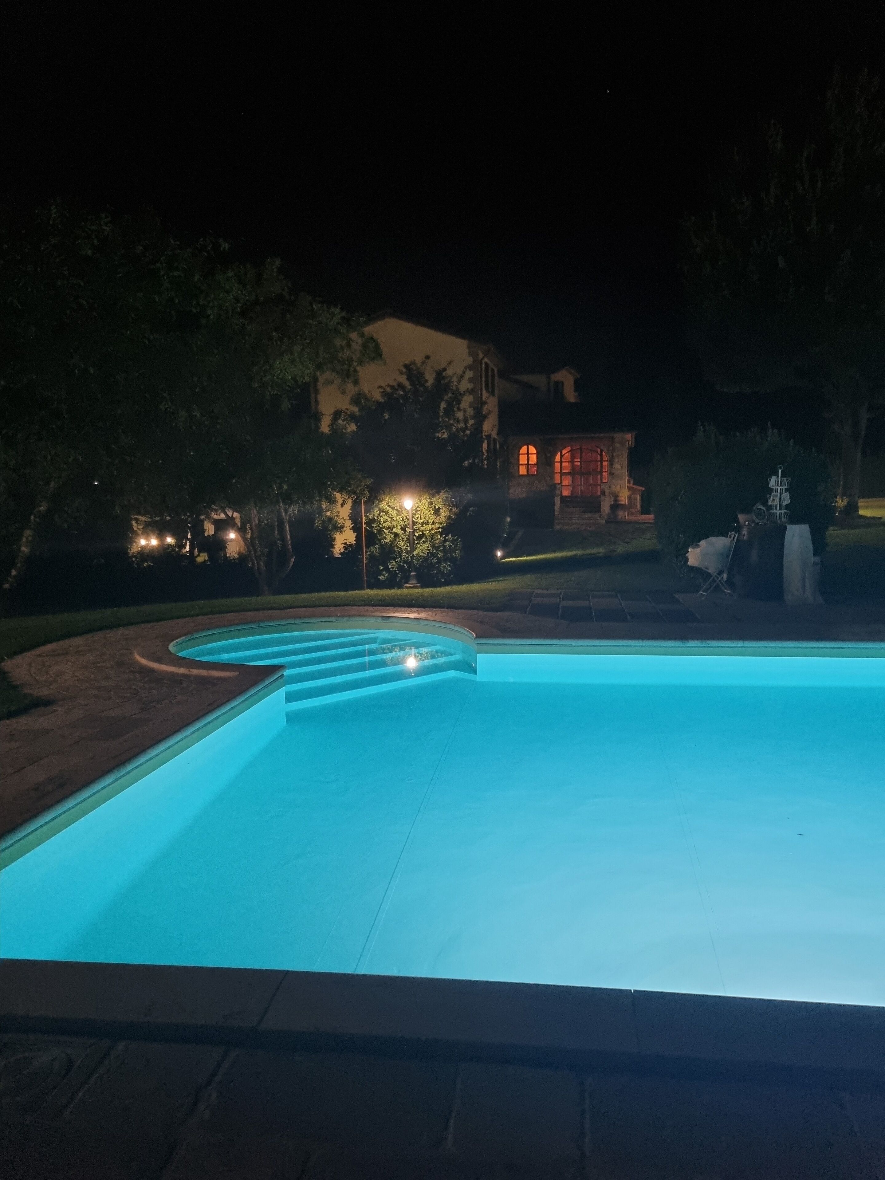 Una piscina iluminada de noche, rodeada de árboles y con un edificio al fondo.