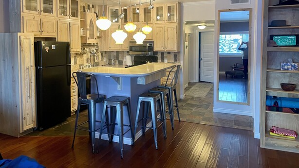Una cucina moderna con mobili in legno, un frigorifero nero, un'isola centrale con sgabelli da bar, illuminazione a sospensione e vista sull'area living adiacente.