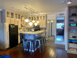 A modern kitchen with wooden cabinets, a black refrigerator, a central island with bar stools, pendant lighting, and a view into the adjacent living area. - Renovated, steps to restaurants, mountain views, on free bus route (Winter Park)