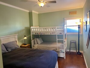 Une chambre avec un lit superposé, un lit simple, une table de chevet avec une lampe, une fenêtre avec des stores et un panneau décoratif mural.