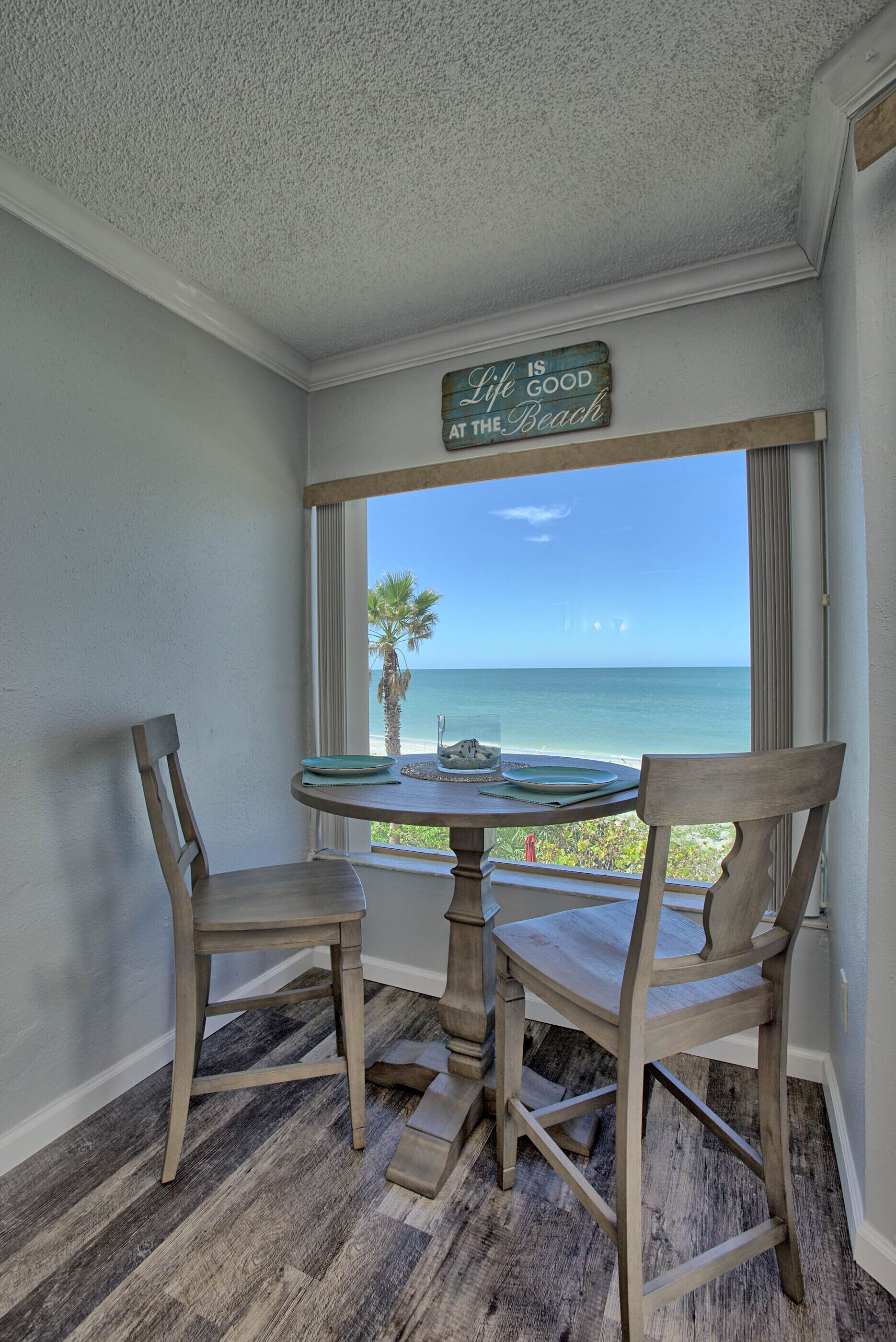 A corner room with a table and two chairs, a window with a view of the beach, and a sign that reads "Life is good at the beach."