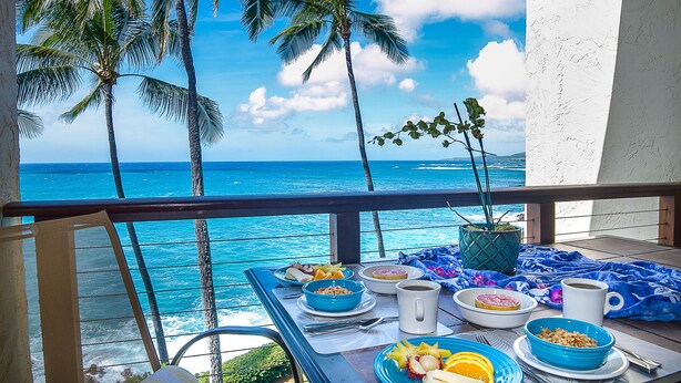 A balcony with a table set for breakfast, overlooking a tropical beach with palm trees and the ocean. - STUNNING OCEANFRONT * Poipu's Best View * New Air Conditioning throughout (koloa)
