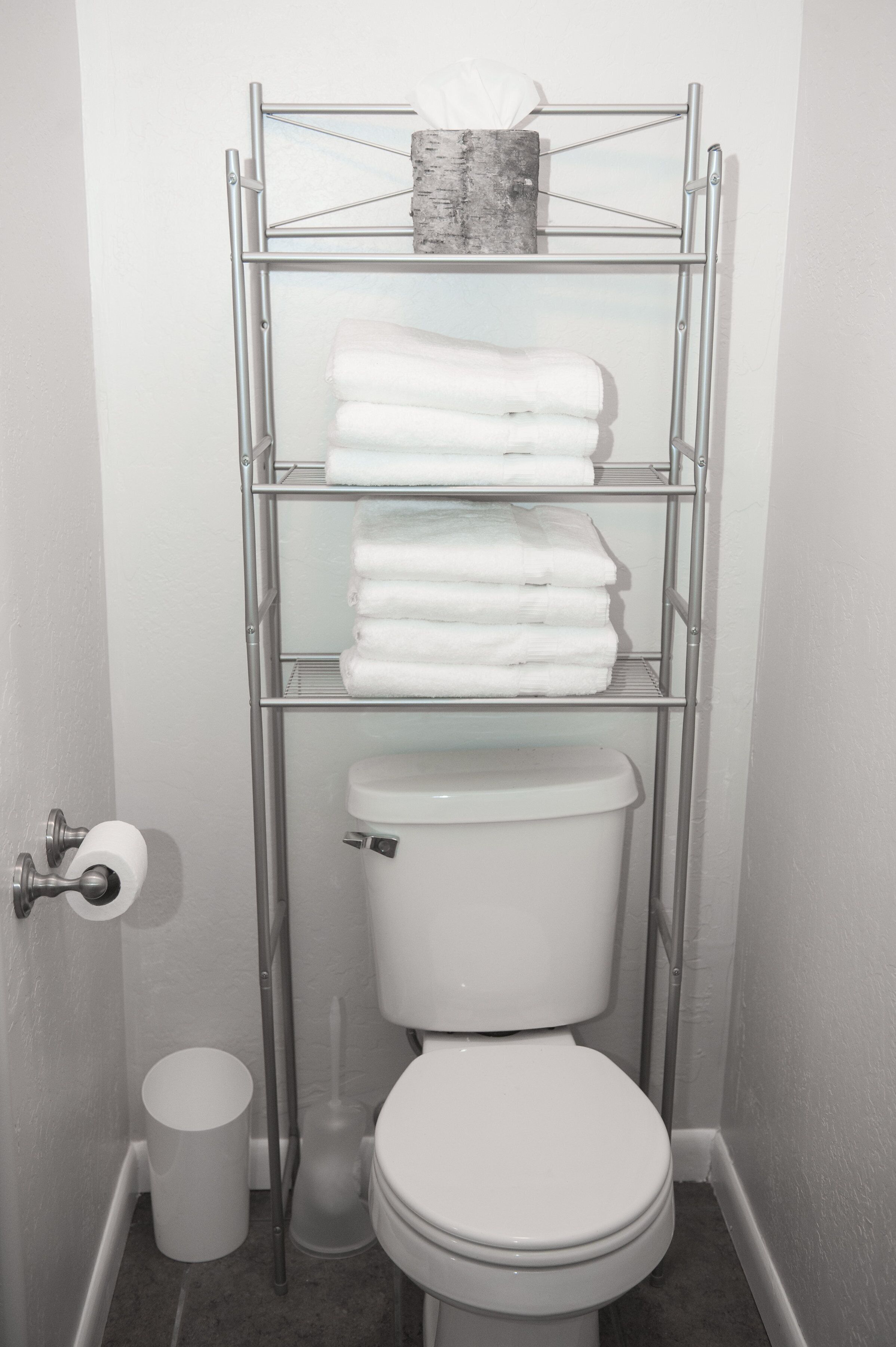 Een badkamer met toilet, een metalen rek met opgevouwen handdoeken, een toiletpapierhouder en een prullenbak.