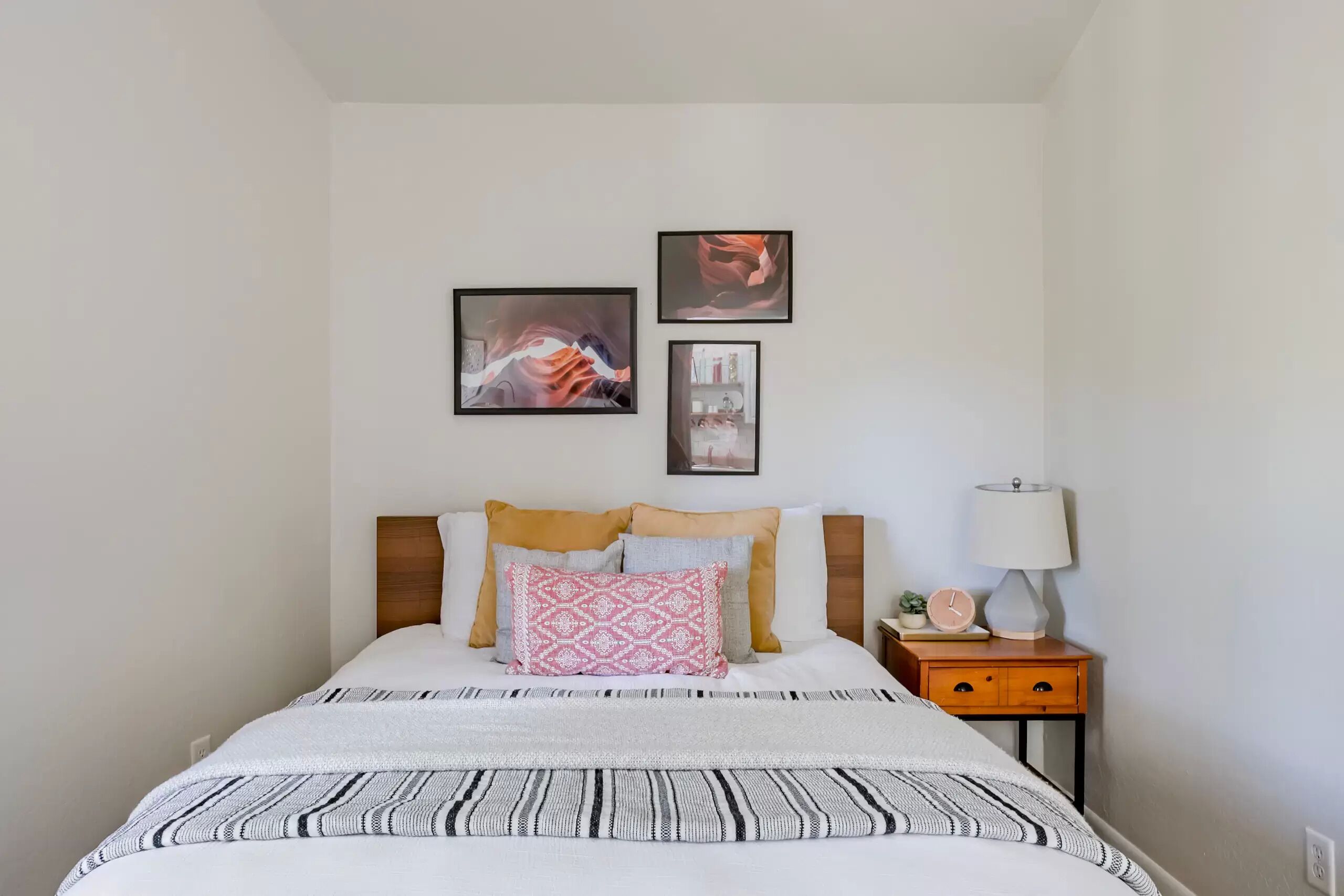 Een net opgemaakt bed met een bedspreidek en kussens, een nachtkastje met een lamp, en drie ingelijste foto's aan de muur.