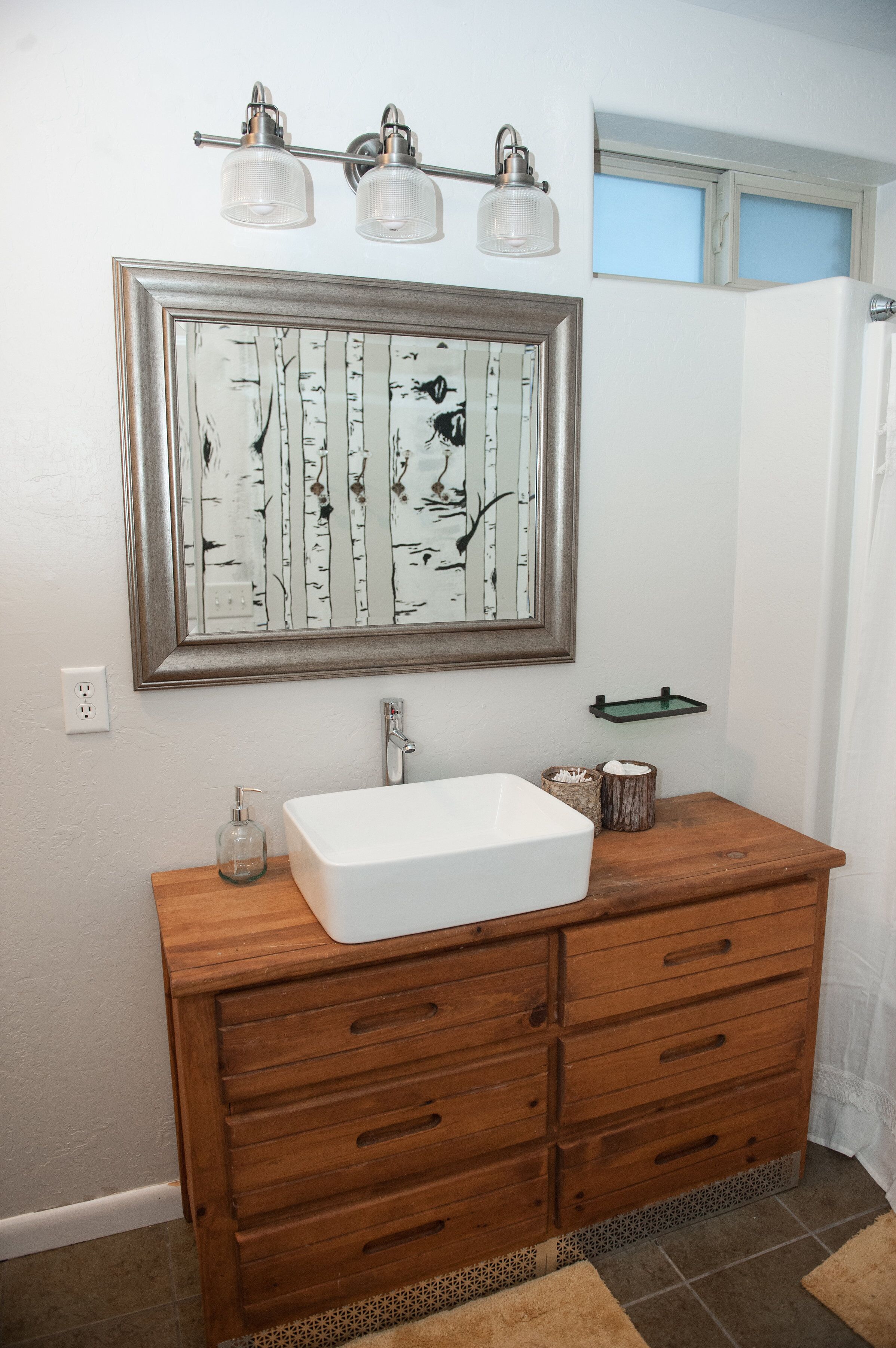 A bathroom with a wooden vanity, a white sink, and a mirror with a birch tree painting.