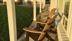 A covered patio area with a wooden chair and a table, a white fence, and a grassy lawn.