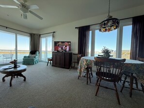 Un salón espacioso con un gran ventanal con vista al mar, un comedor con mesa y sillas, un televisor y un ventilador de techo.