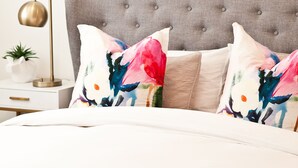 Una cama bien hecha con cabecera tapizada gris, un cuadro abstracto enmarcado encima y una mesita de noche con una lámpara y una planta en maceta.