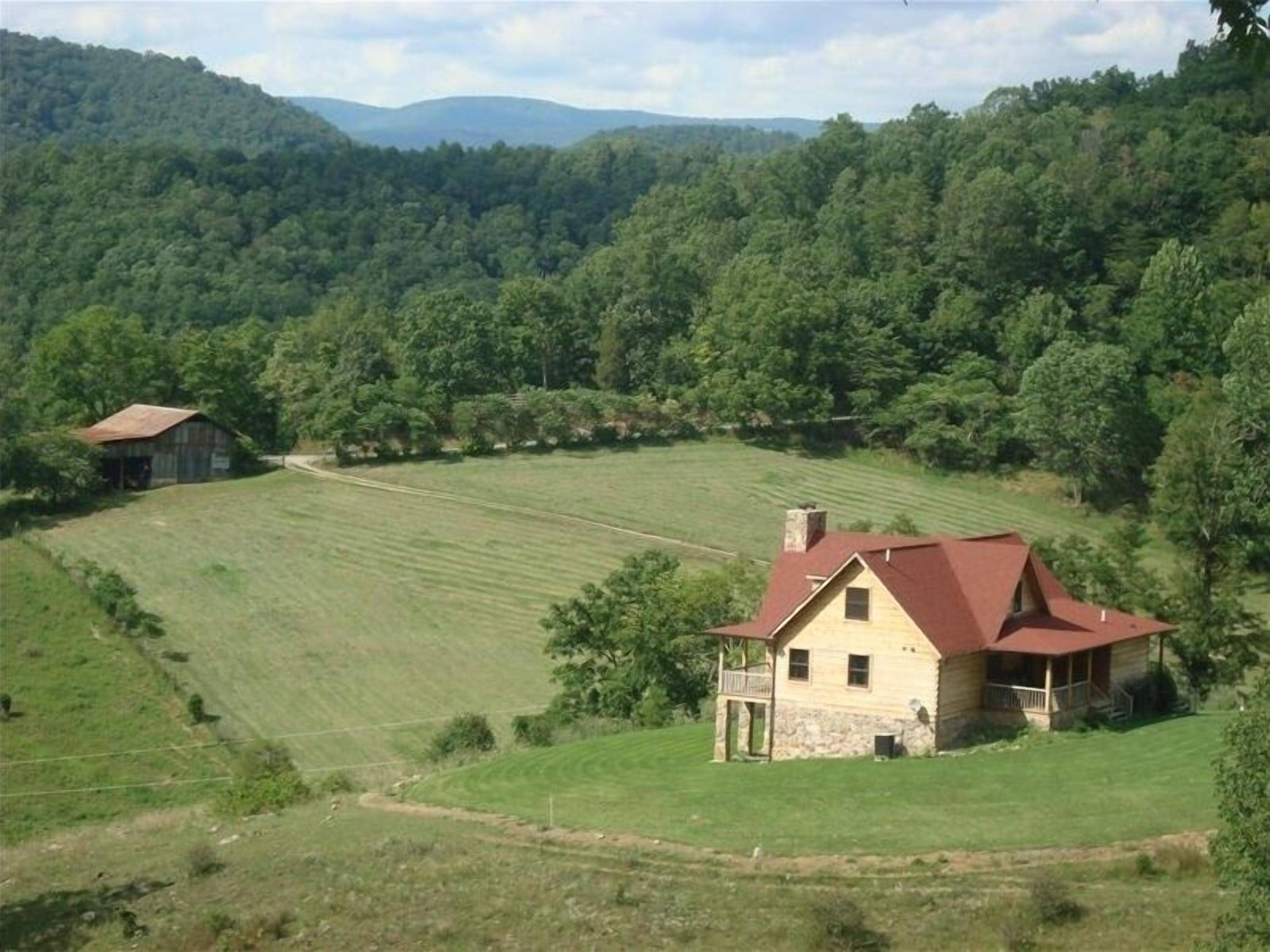 A rural house with a red roof, surrounded by a green field and forested hills.