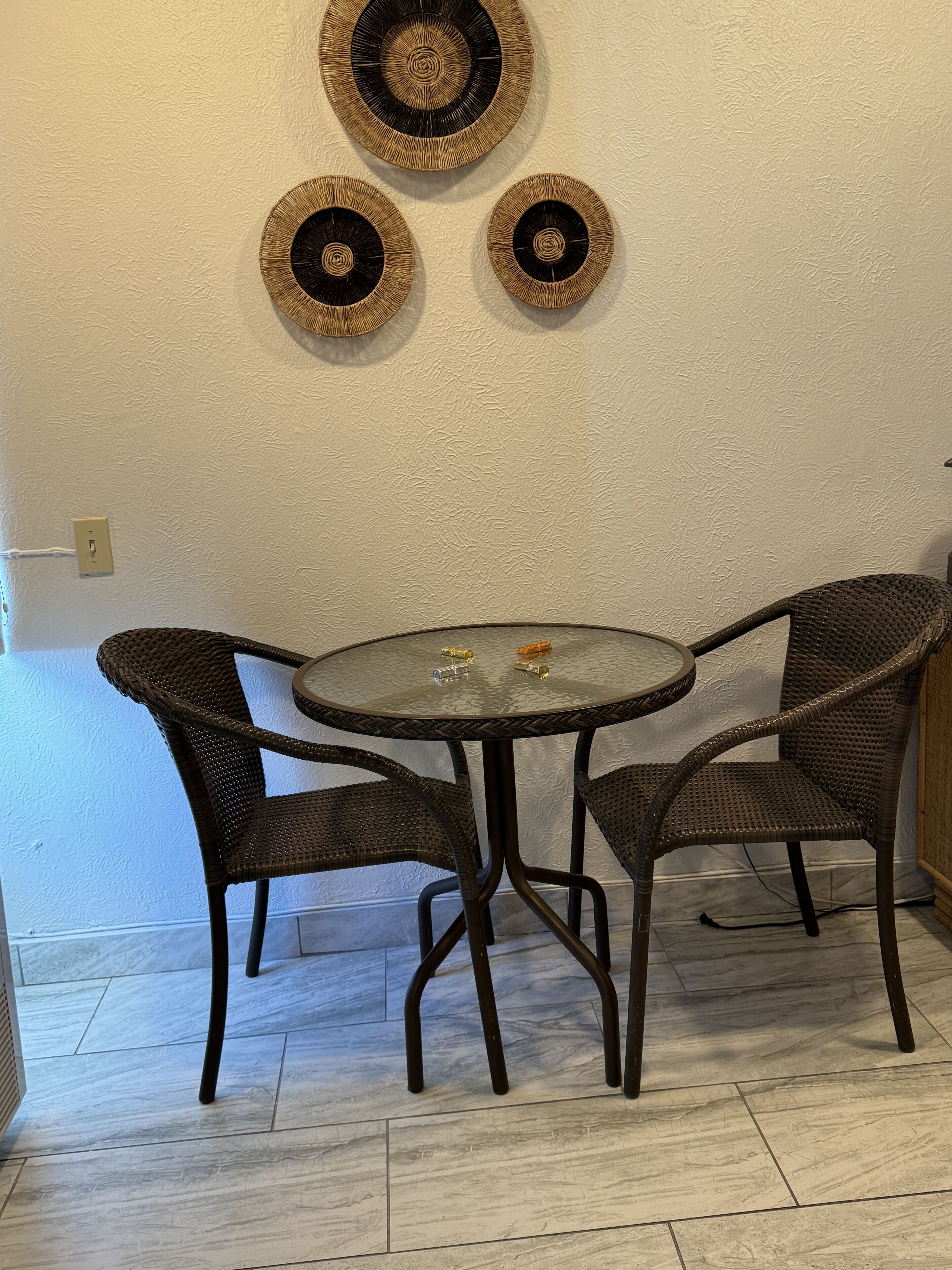 A dining area with a round table and four wicker chairs, a textured wall, and a light switch.