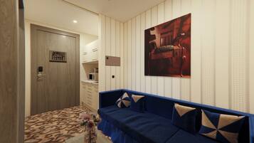 Quarto de hotel com porta de madeira, cozinha compacta, sofá azul com almofadas estampadas e uma obra de arte emoldurada na parede.