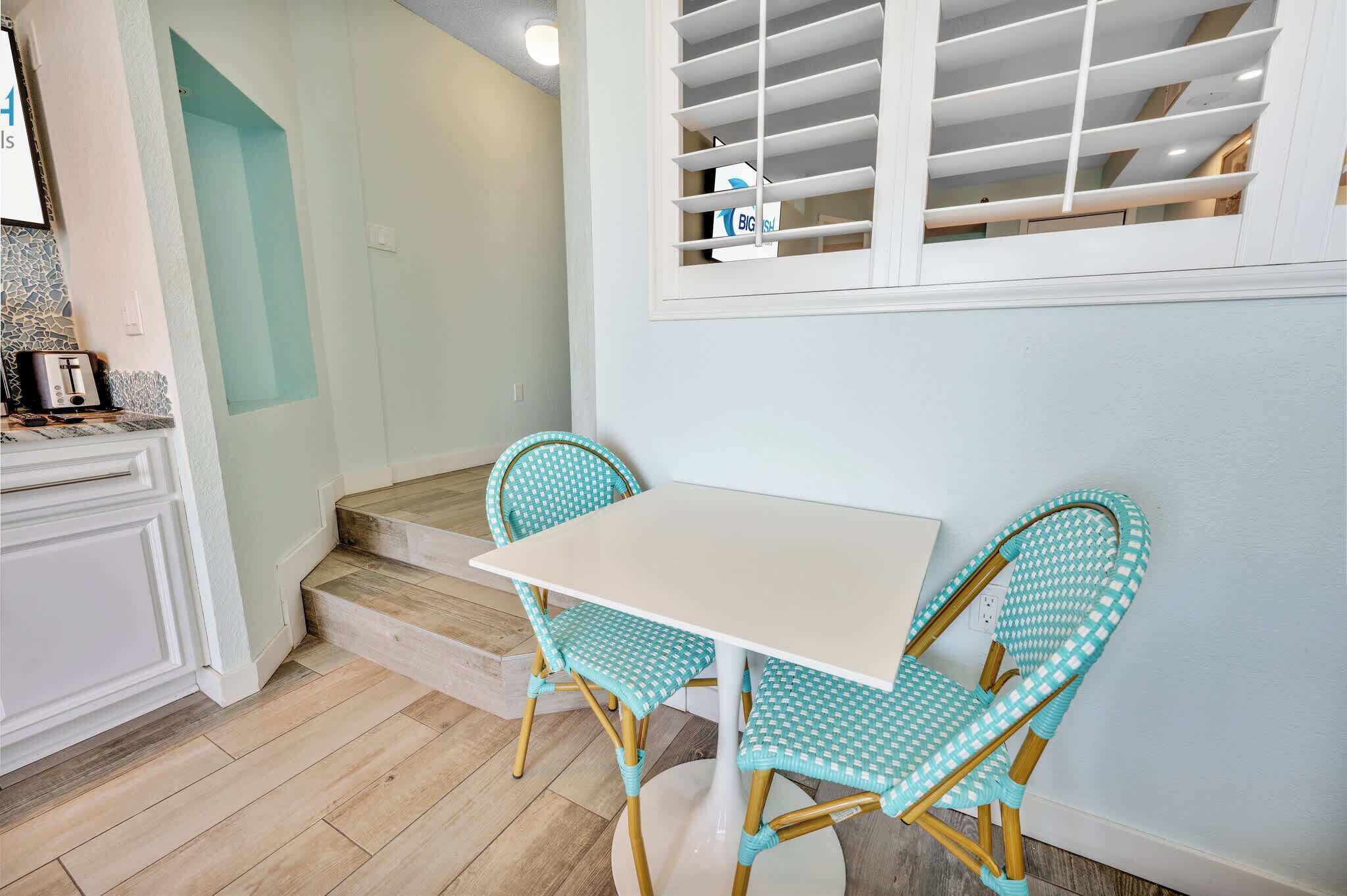 Una cocina con una mesa blanca y dos sillas turquesas, una ventana con persianas y una escalera que lleva a otra habitación.