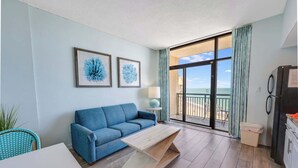 Un salon avec un canapé bleu, une table basse en bois et une table à manger avec des chaises. Deux tableaux encadrés ornent le mur, ainsi qu’une lampe. Une porte coulissante offre une vue sur l’océan.