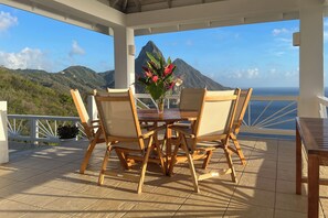 A wooden table with four chairs and a vase of flowers on a covered patio overlooking a mountainous landscape.