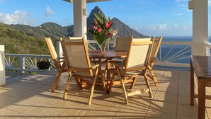 A covered outdoor area with a table and chairs, a vase of flowers, and a view of mountains and the ocean.