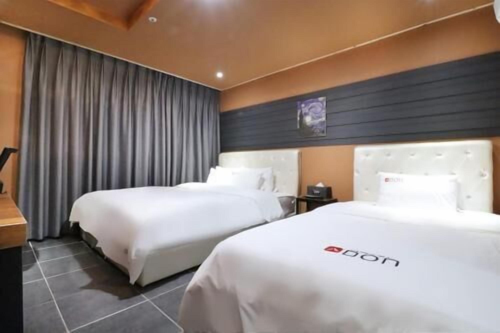 A hotel room with two beds, a large window with curtains, a wooden headboard, and a wall-mounted artwork.