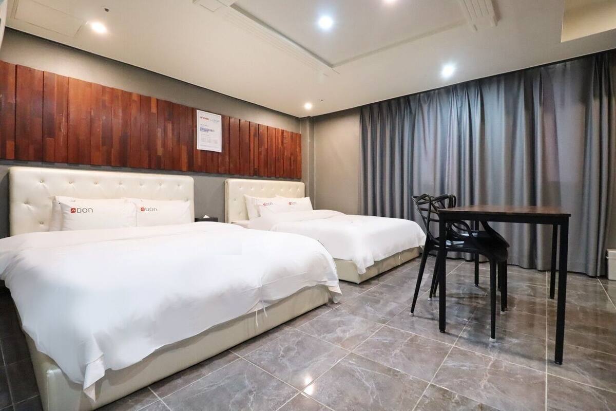 Ein Hotelzimmer mit zwei Betten, einem Holzkopfteil, einem Esstisch mit Stühlen und einem Vorhang.
