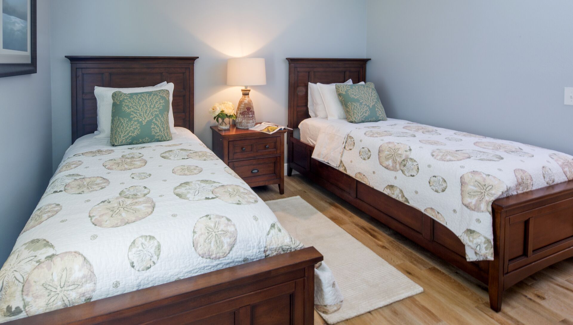 A bedroom with two wooden beds, each with a floral-patterned bedspread, a nightstand with a lamp, and a small table with a vase of flowers.