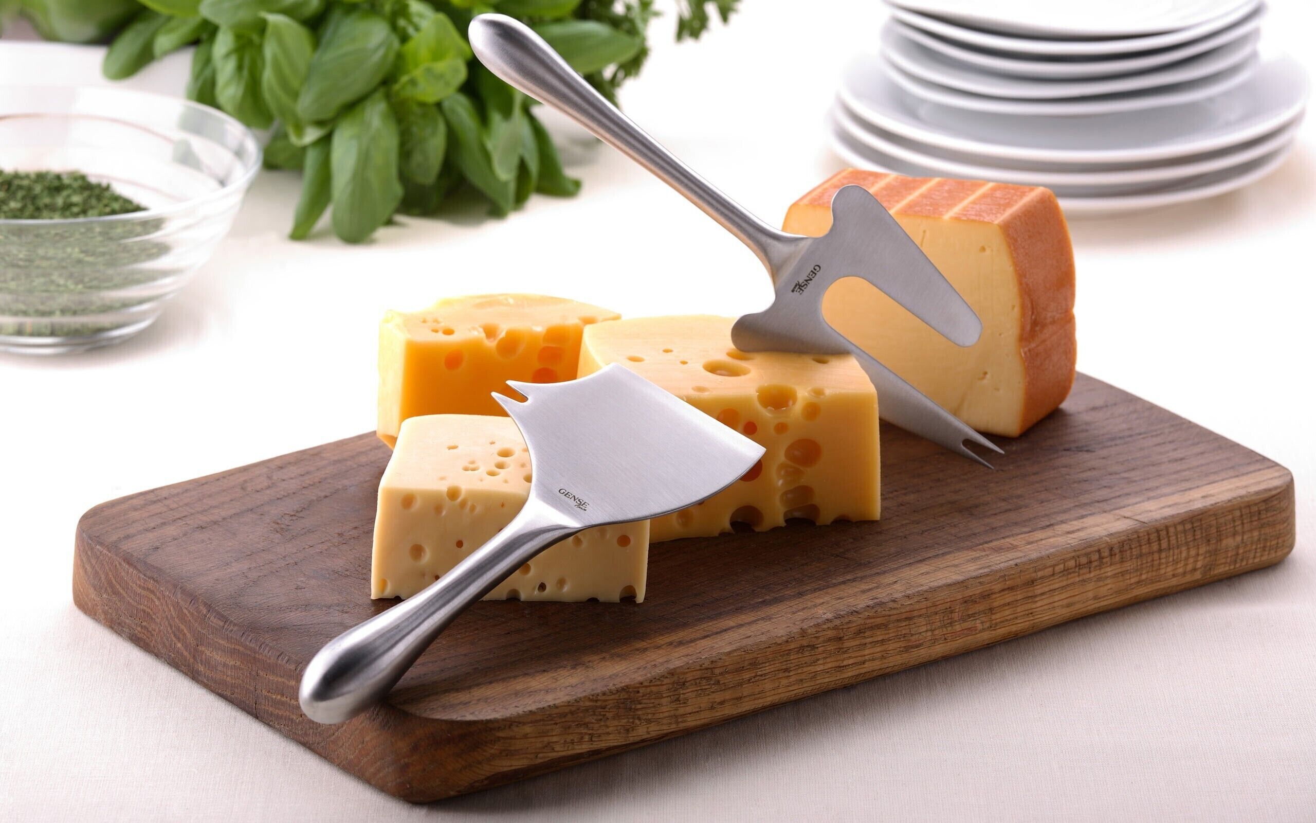A wooden cutting board with a cheese slicer, a wedge of cheese, and a bowl of green herbs.
