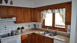A kitchen with wooden cabinets, a stove, a sink, and a window with a floral curtain. - Lovely cottage in West Asheville. Hot Tub. Pool open for SUMMER! (Asheville)