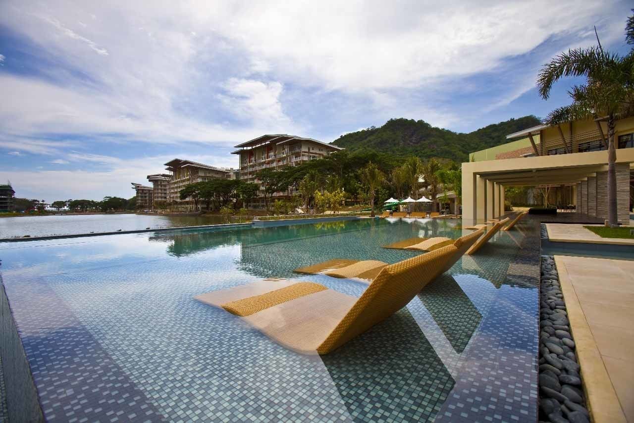A swimming pool with lounge chairs, surrounded by buildings and a mountainous backdrop.